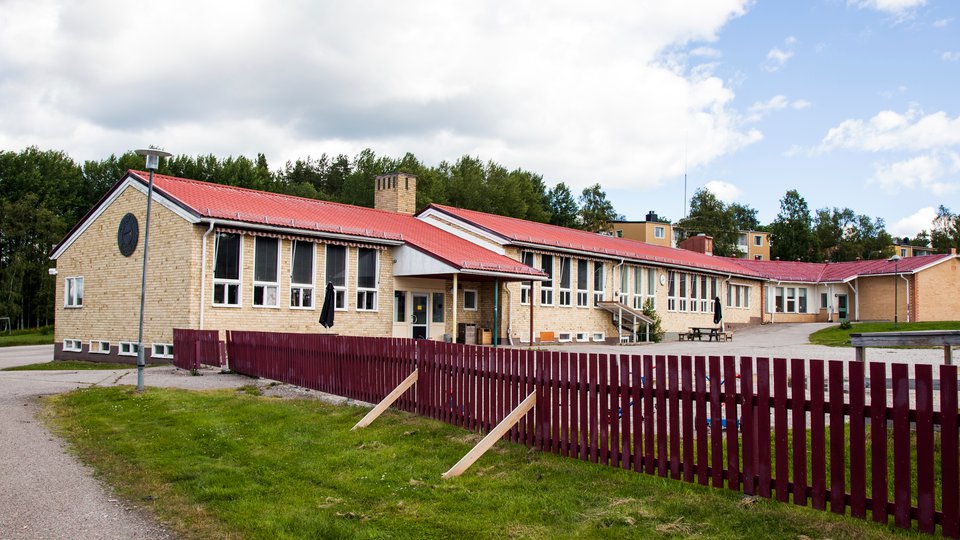 En ljusgul tegelbyggnad med ett rött staket i förgrunden. Ilsbo skola.