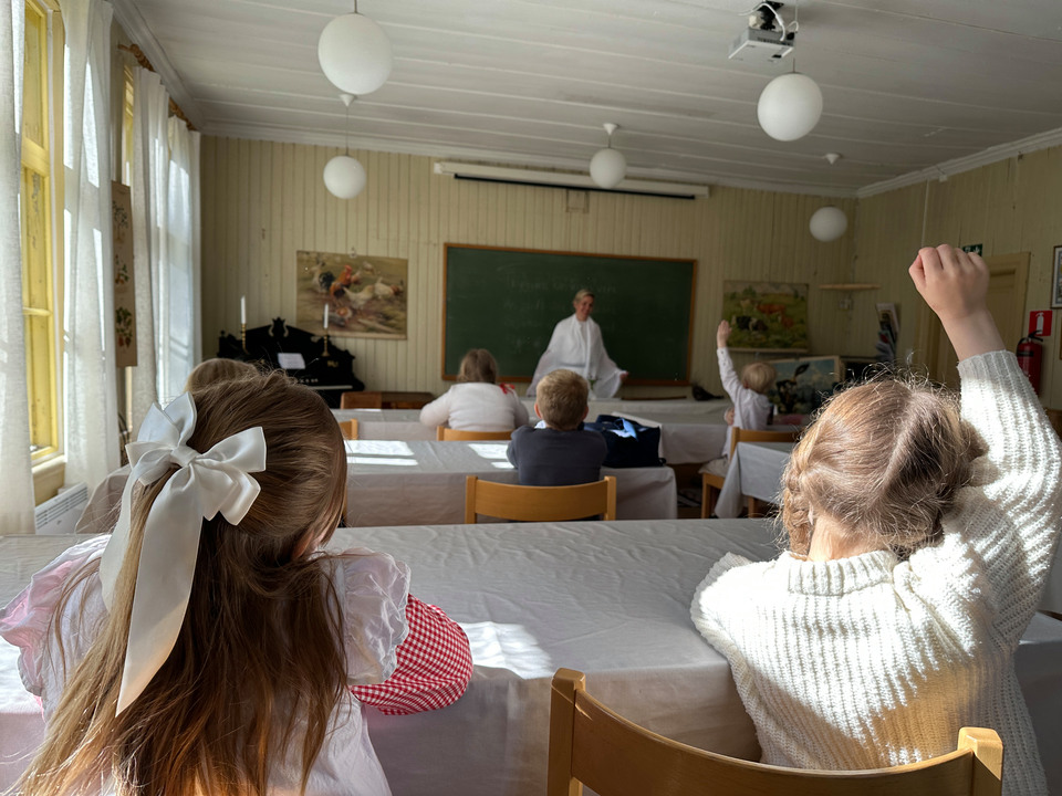 Elever sitter i ett glassrum under en lektion. Längst fram vid svarta tavlan står en kvinnlig lärare. Flera elever räcker upp handen.