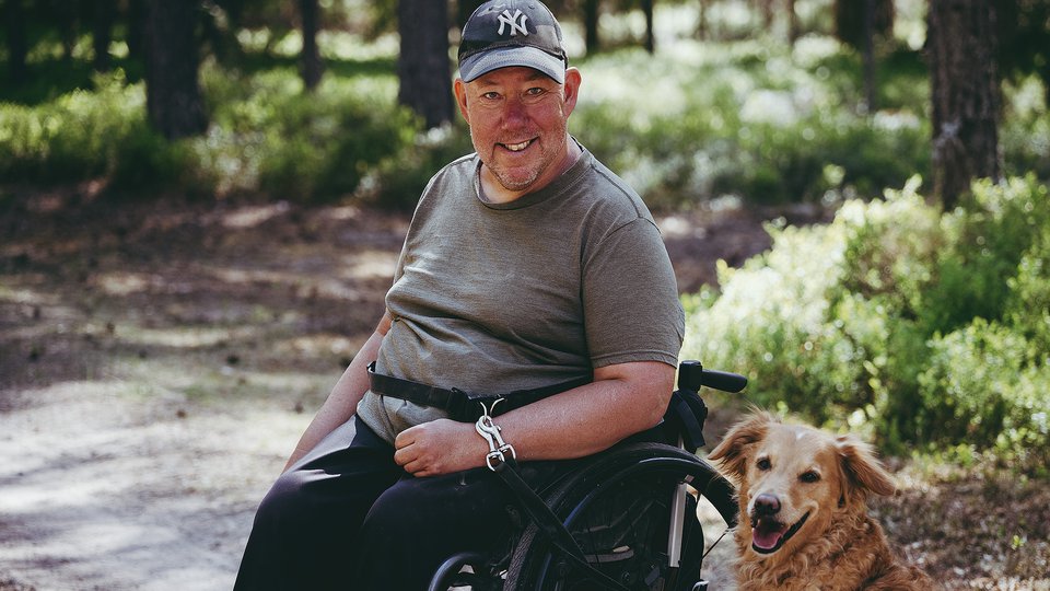 En leende man sitter i en rullstol på en grusväg i skogen. Intill honom sitter en hund.