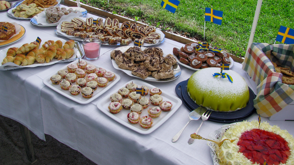 Ett bord fullt av tårtor, fikabröd och Sverigeflaggor utomhus i solen.