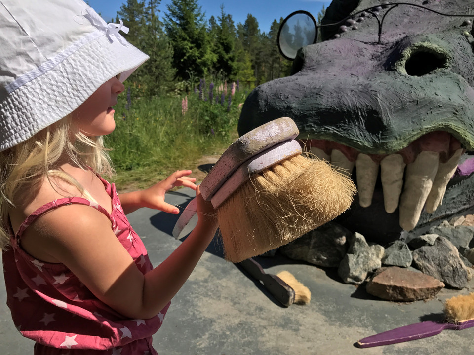 Flicka med solhatt och med en stor tandborste i handen framför en stor drakes framtänder.