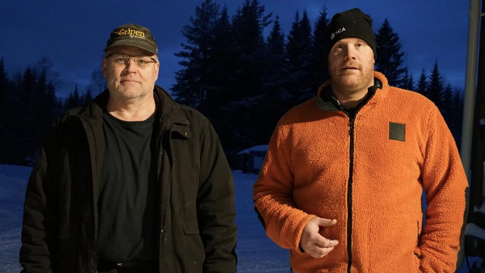 Två män utomhus med snö och skog i bakgrunden.
