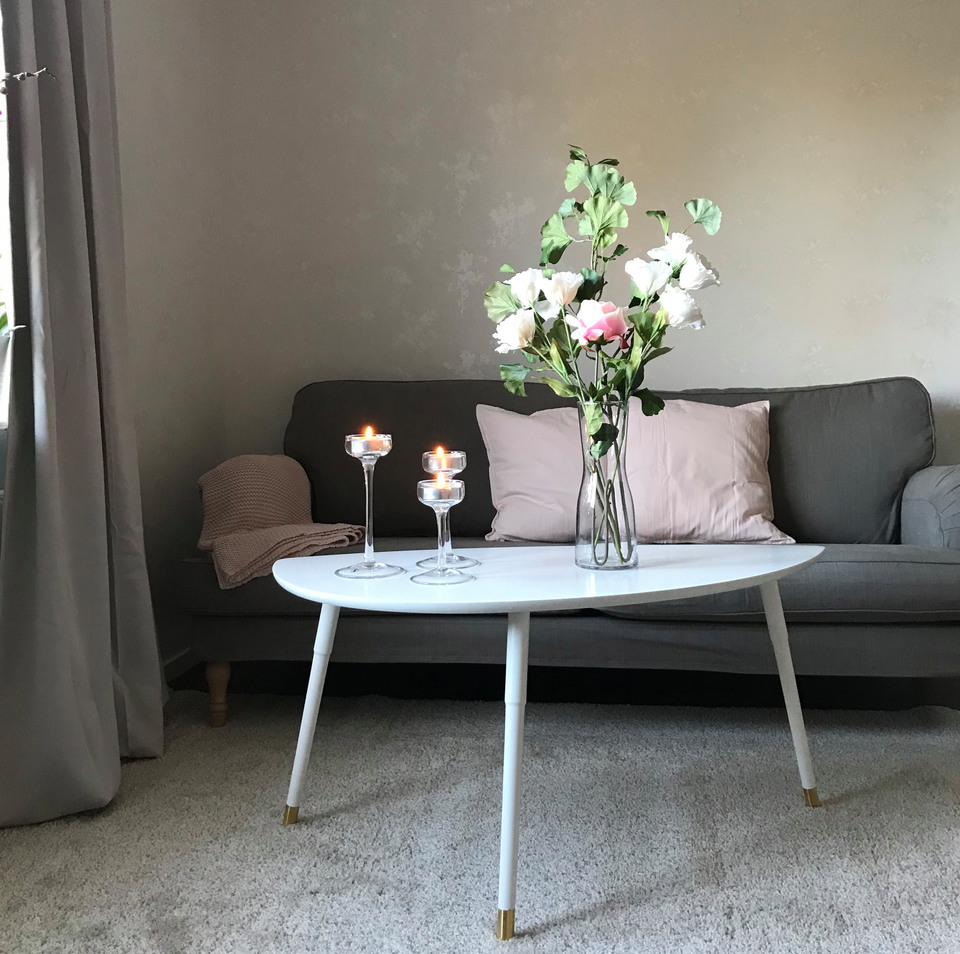 Ett vardagsrum med en grå soffa och ett vitt bord. På bordet finns en vas med blommor och bredvid står två ljusstakar med tända ljus.