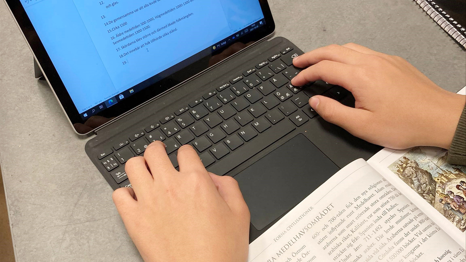 Ett barns händer knappar på en elevdator med en lärobok uppslagen på skolbänken.