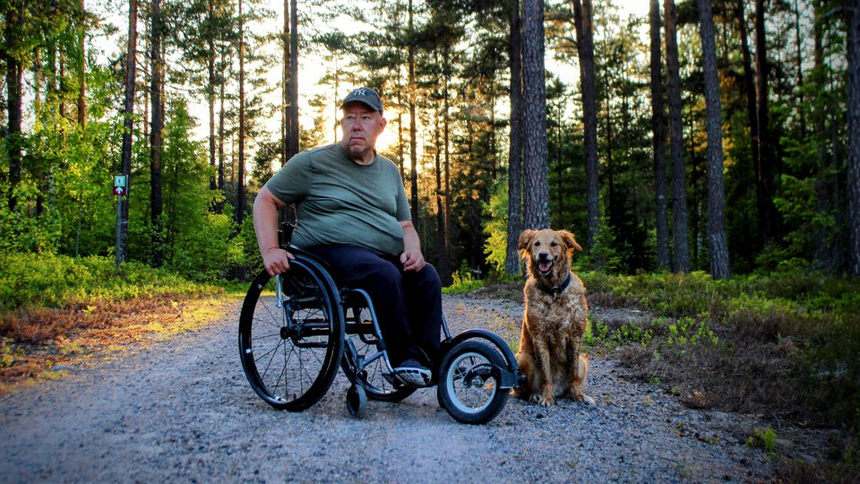En man sitter i en rullstol på en grusväg i skogen. Intill honom sitter en hund.
