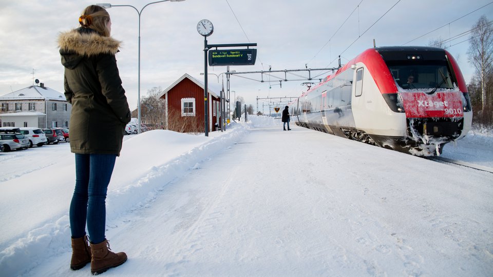 En kvinna står vid ett litet rött stationshus och ett regionaltåg.