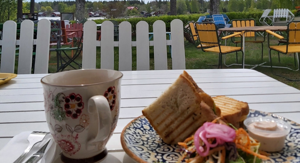 Ett kafébord utomhus  med en tekopp och ett fat med en varm smörgås på.