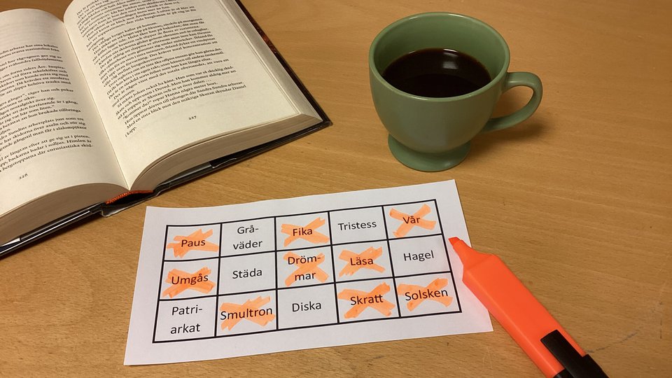 En uppslagen bok, en kopp kaffe och en bingobricka med ord. Intill bingobrickan ligger en överstrykningspenna.