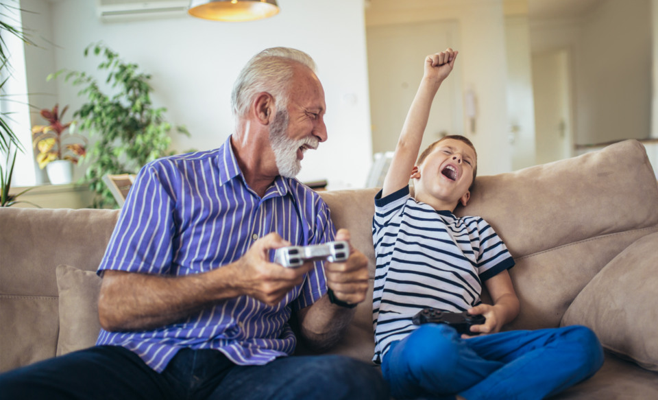 En äldre man sitter i en soffa med sitt barnbarn och spelar tv-spel.