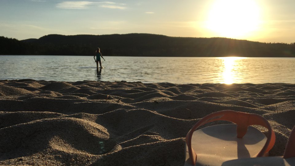 Iförgrunden syns ett par sommarsandaler i på en sandstrand, i bakgrunden syns en person som badar i solnedgången.