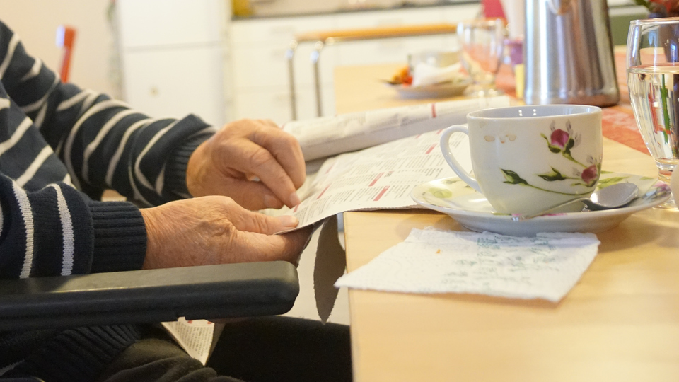 Händer på en äldre man håller i en tidning som ligger på ett bord med en kaffekopp 