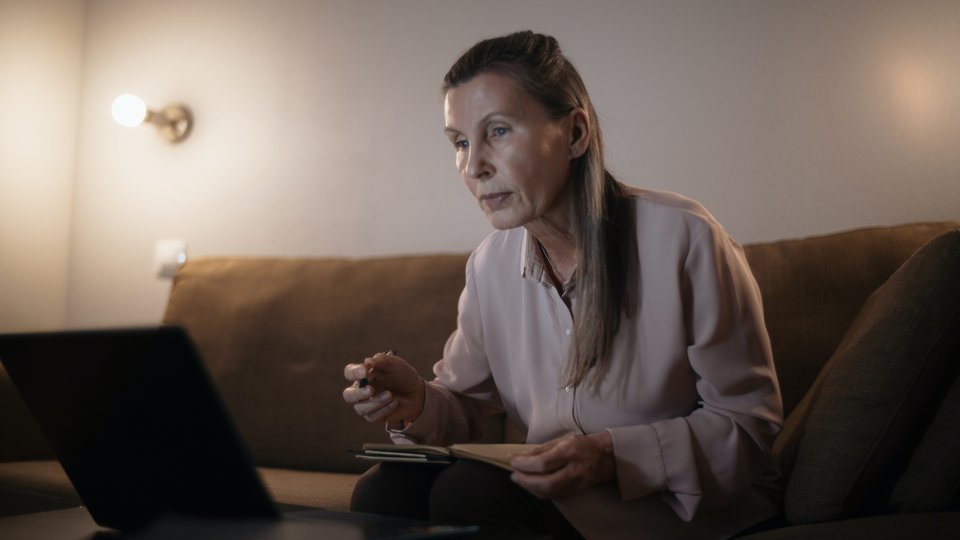 En kvinna sitter i en soffa med papper och penna och tittar på en dataskärm.